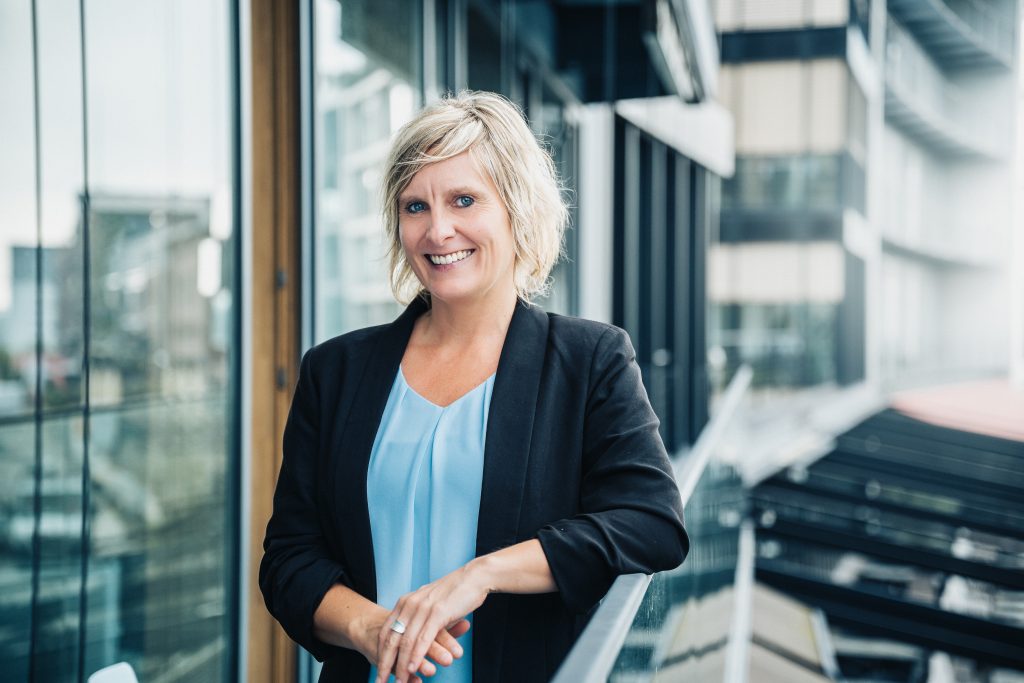 Verena Arnhold mit hellblauem Oberteil und dunklem Blaser draußen vor einer Glasfassade mit anderen Gebäuden im Hintergrund